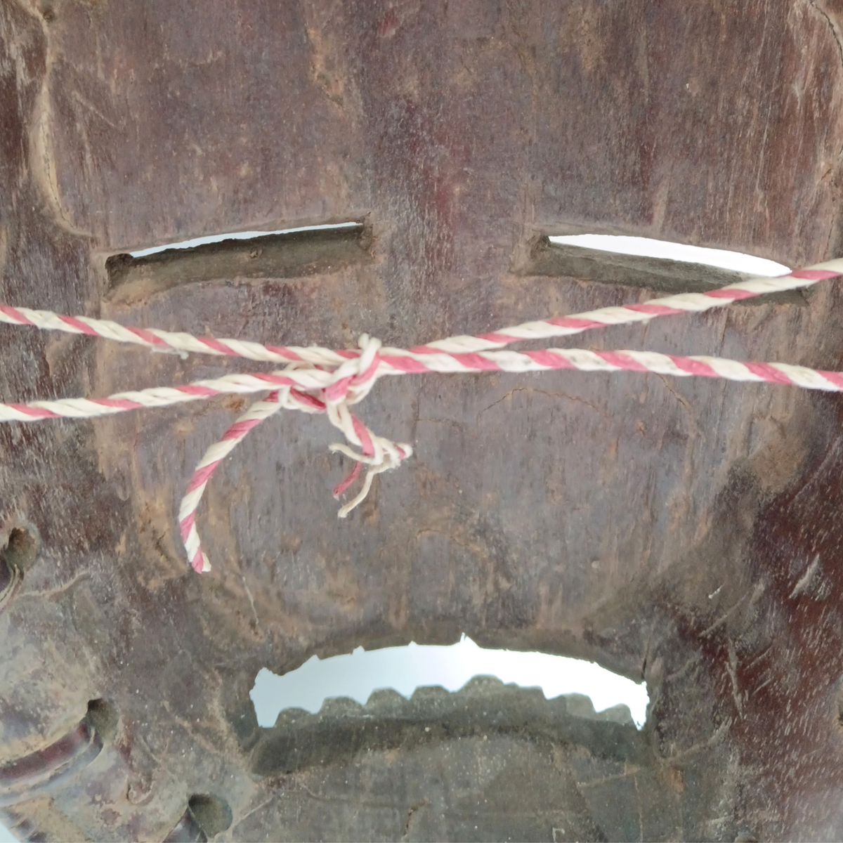 A close-up of a dark carved wooden mask with cutout facial features, tied across the centre with a red-and-white striped rope.