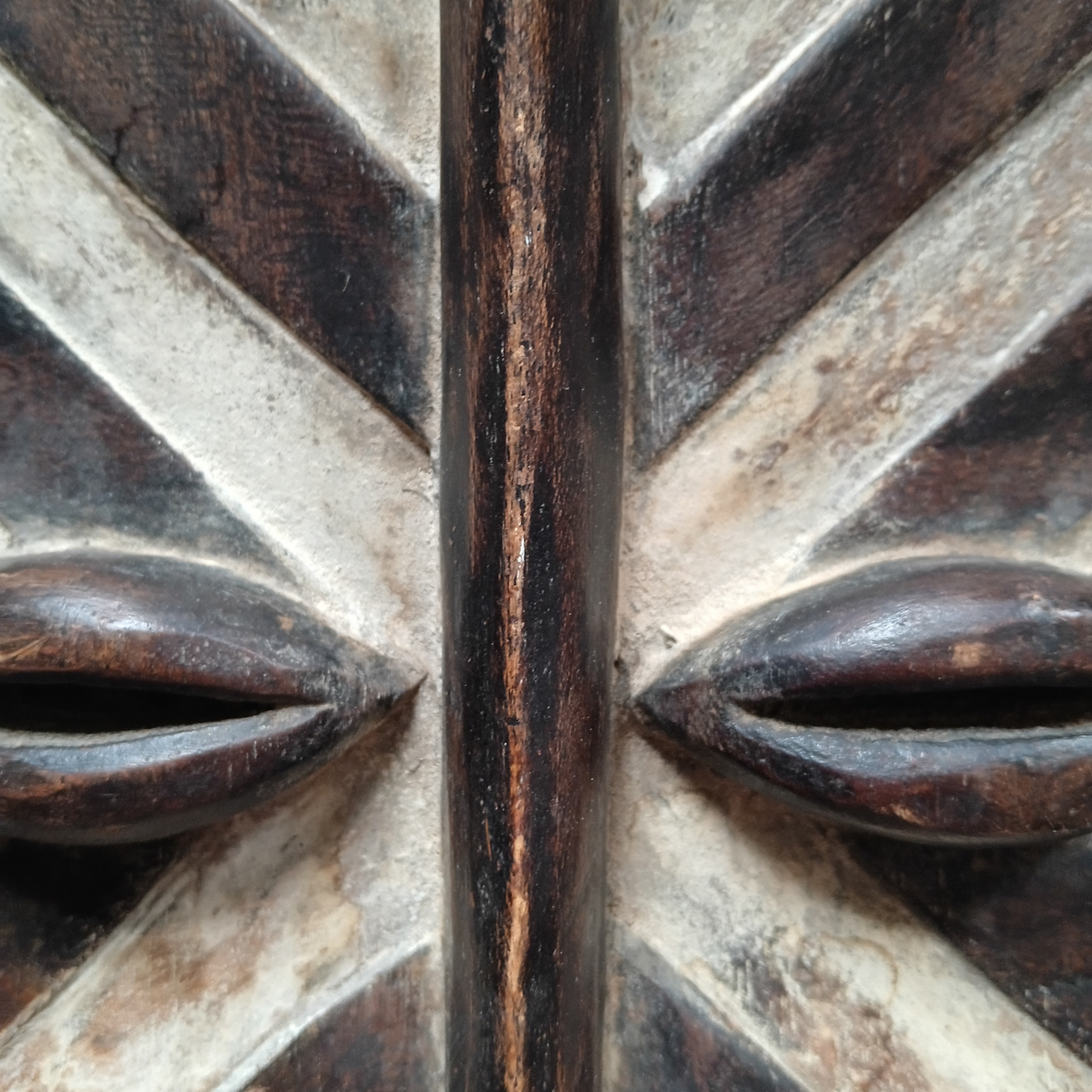 Close-up of a carved wooden mask with two carved eye shapes and radiating carved lines. 