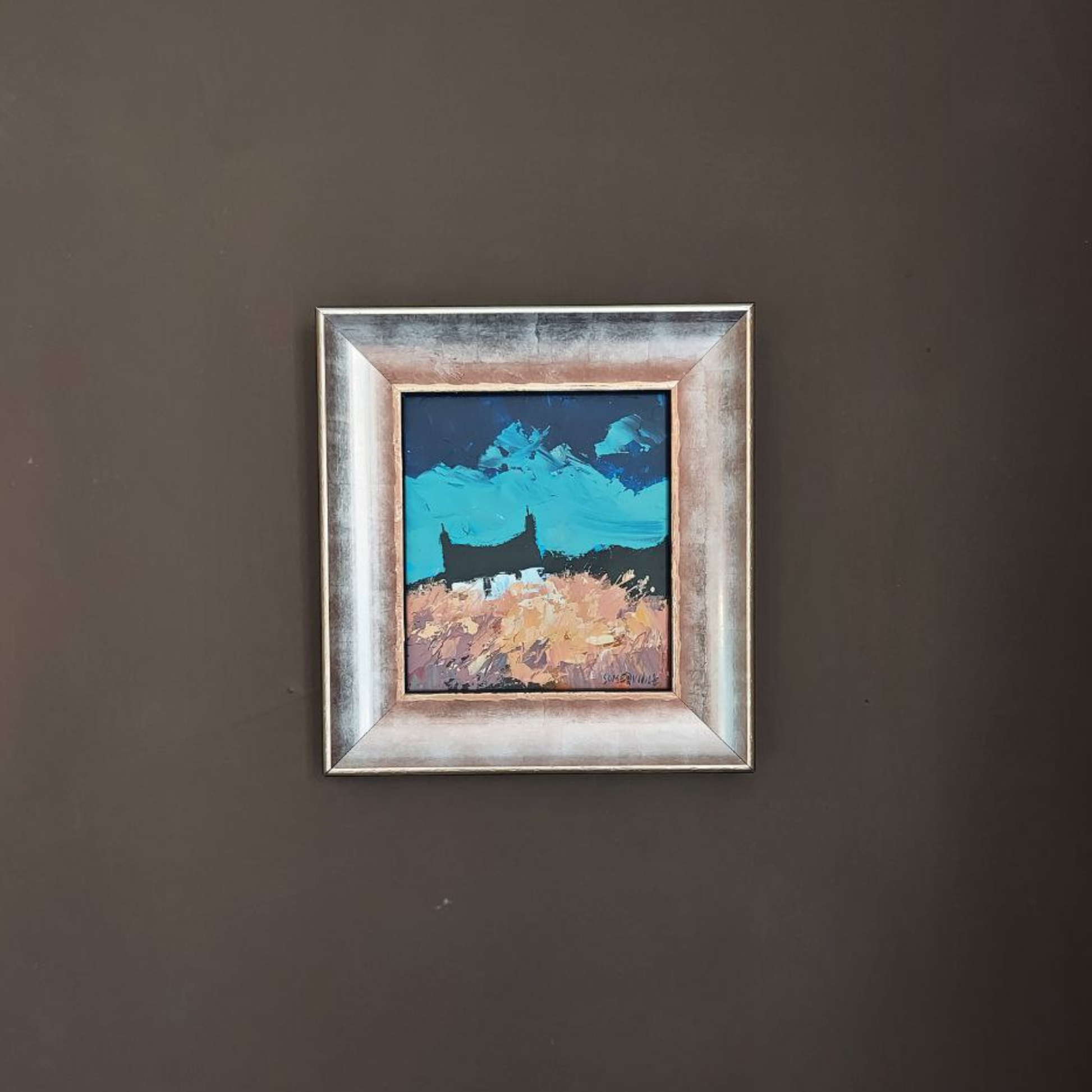 Framed George Somerville artwork of a landscape with mountains and a house on a dark wall.