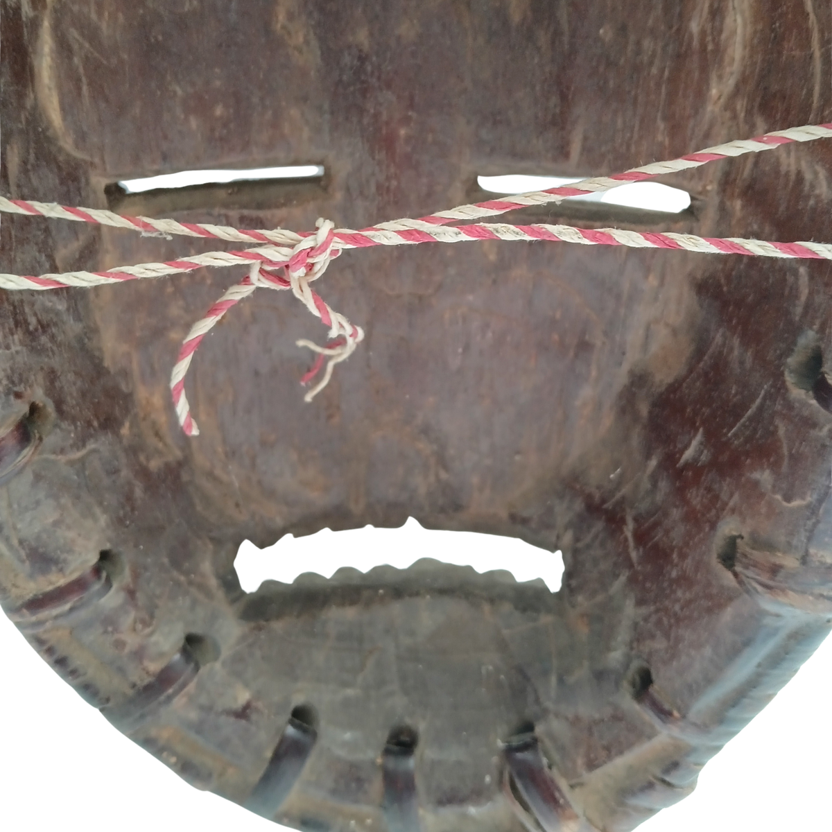 A close-up of a dark brown wooden mask with cutouts, secured by a red and white twisted string tied around the centre, against a plain white background.