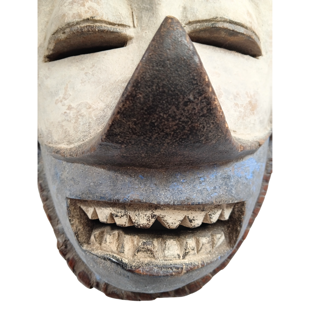 A close-up of a carved wooden mask with a wide, toothy grin, a dark triangular nose, and weathered beige and blue-grey painted details on a white background.
