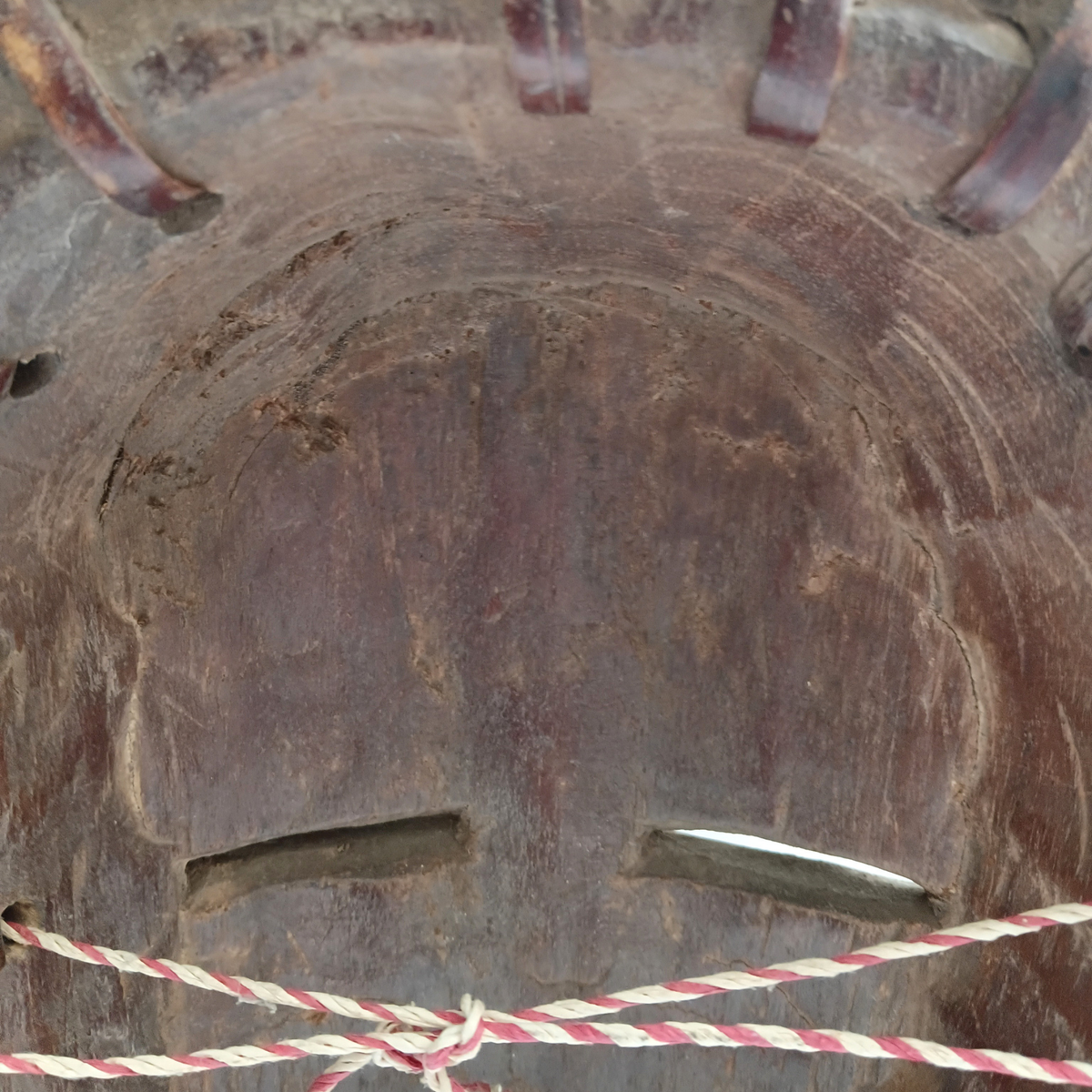 Close-up of a dark brown carved wooden mask with carved facial features, small cutouts, and red-and-white rope tied across the front.