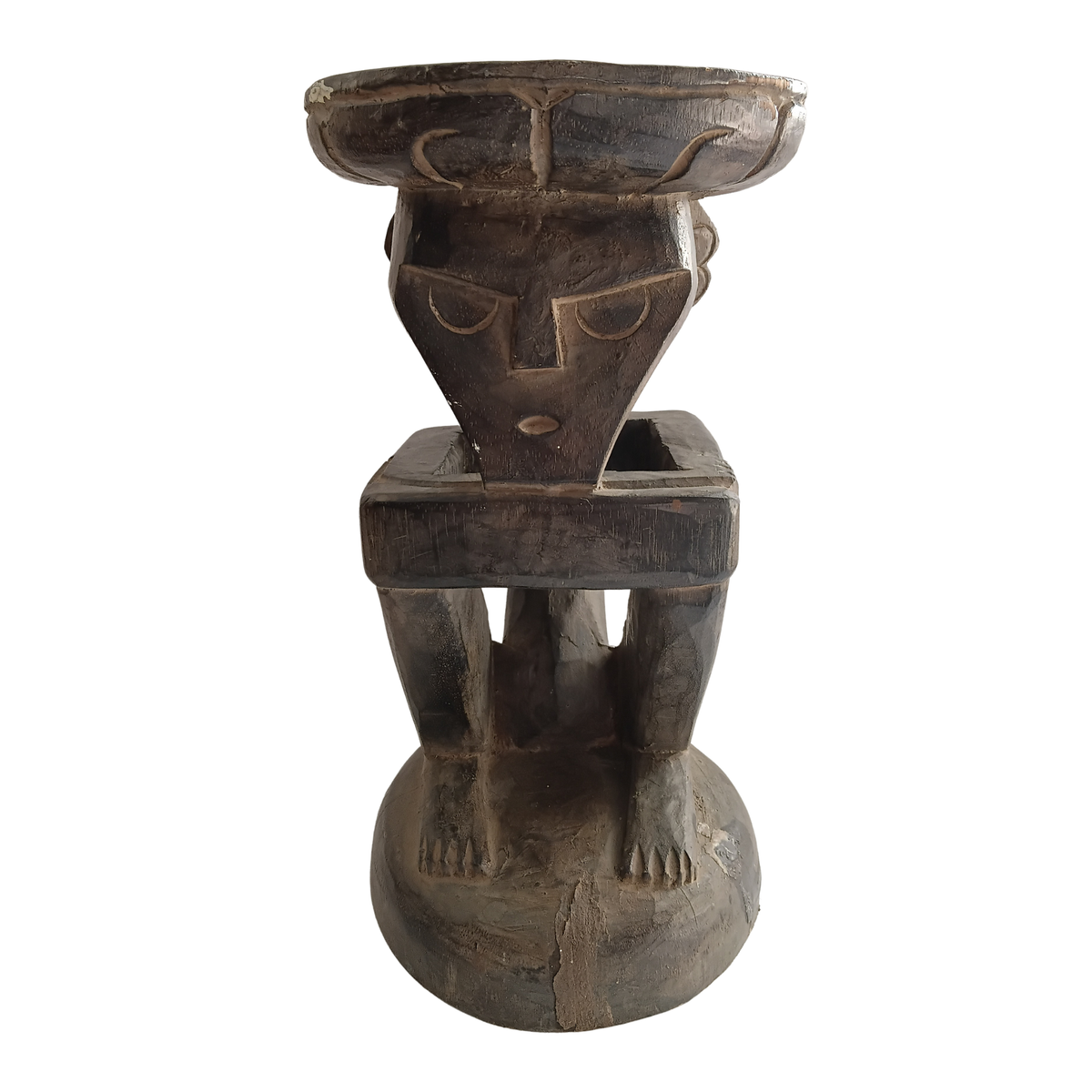A dark carved wooden pedestal sculpture with a stylized human face and legs, photographed against a plain white background.