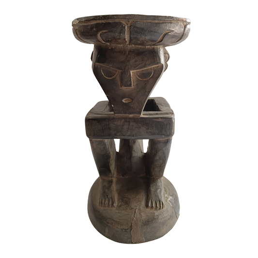 A dark carved wooden pedestal sculpture with a stylized human face and legs, photographed against a plain white background.