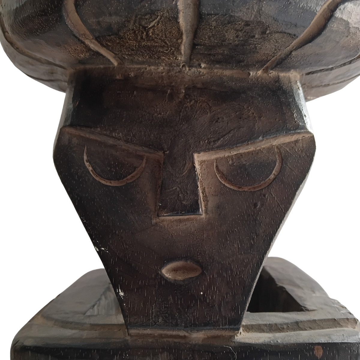 Close-up of a dark carved wooden sculpture or decorative object with a stylized face and textured surface, shown against a plain white background.