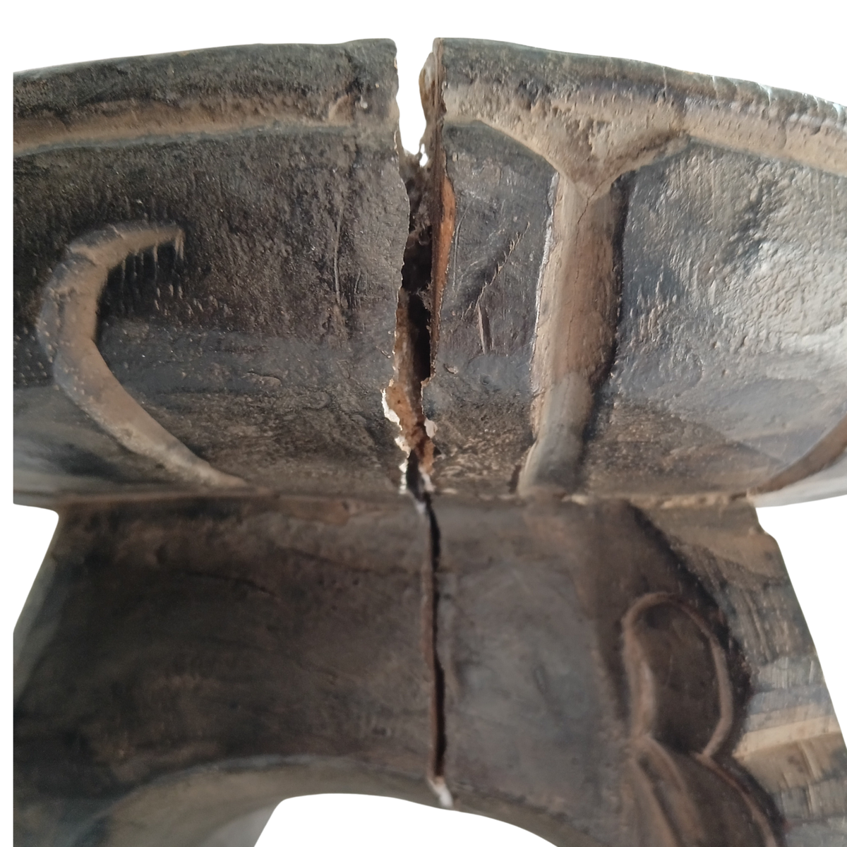 Close-up of a carved wooden stool with a prominent vertical crack running down the centre, showing weathered dark brown wood grain and carved details on both sides against a white background.