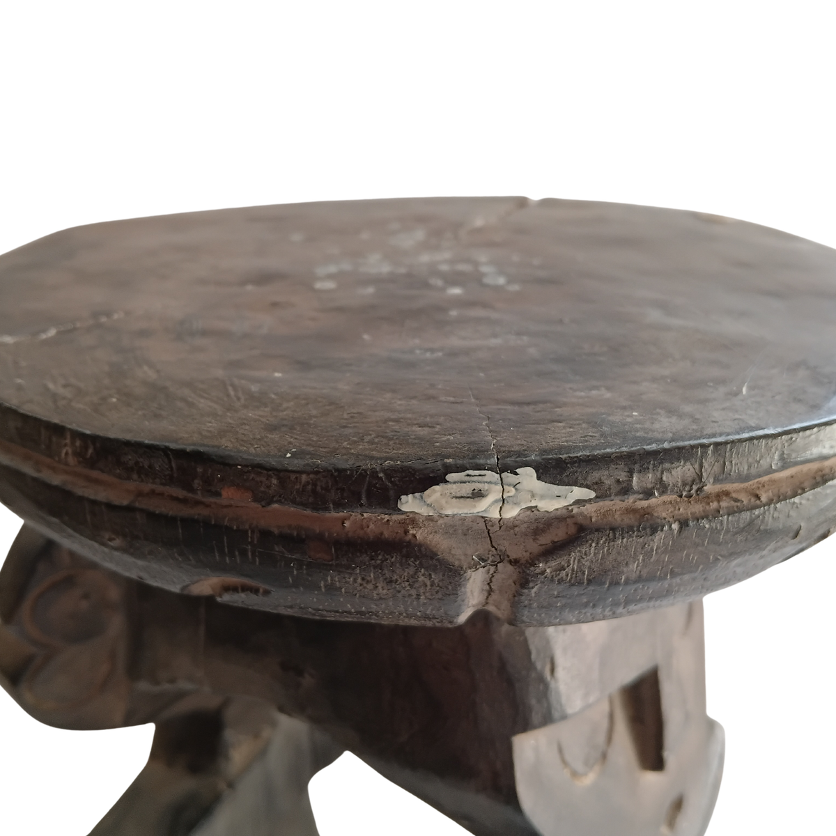 A close-up of a dark, distressed wooden stool with a circular top and carved legs, showing signs of age and wear against a plain white background.