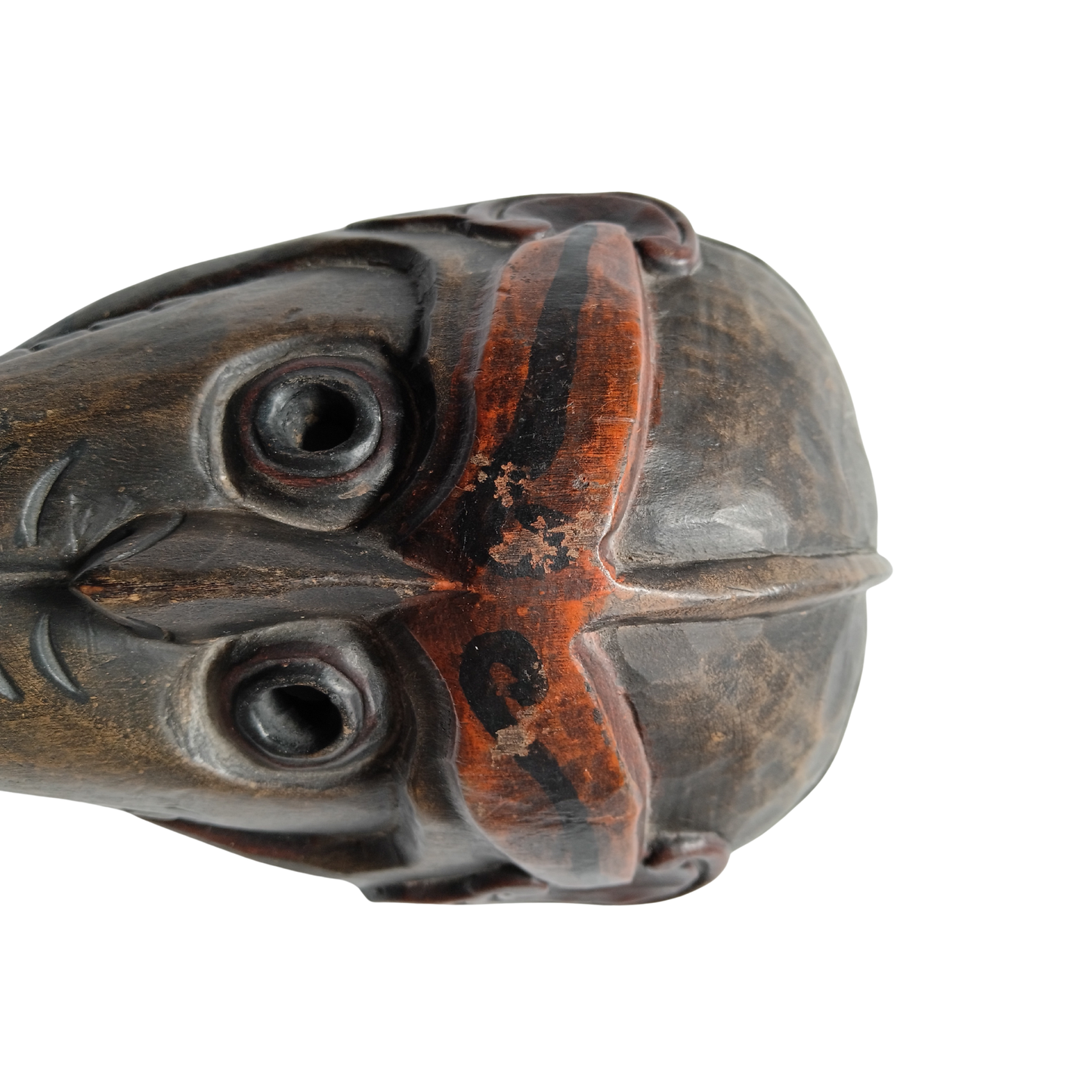 A close-up of a dark carved wooden object, showing detailed hand-hewn textures, a circular eye-like carving, and a prominent reddish-brown painted accent on a plain white background.