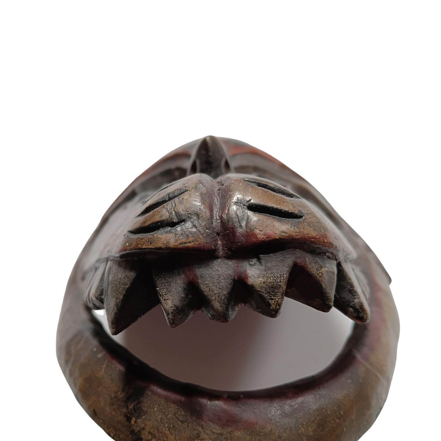 A close-up of a dark brown carved wooden mask or decorative object with a stylized face and sharp teeth-like protrusions, shown against a plain white background.