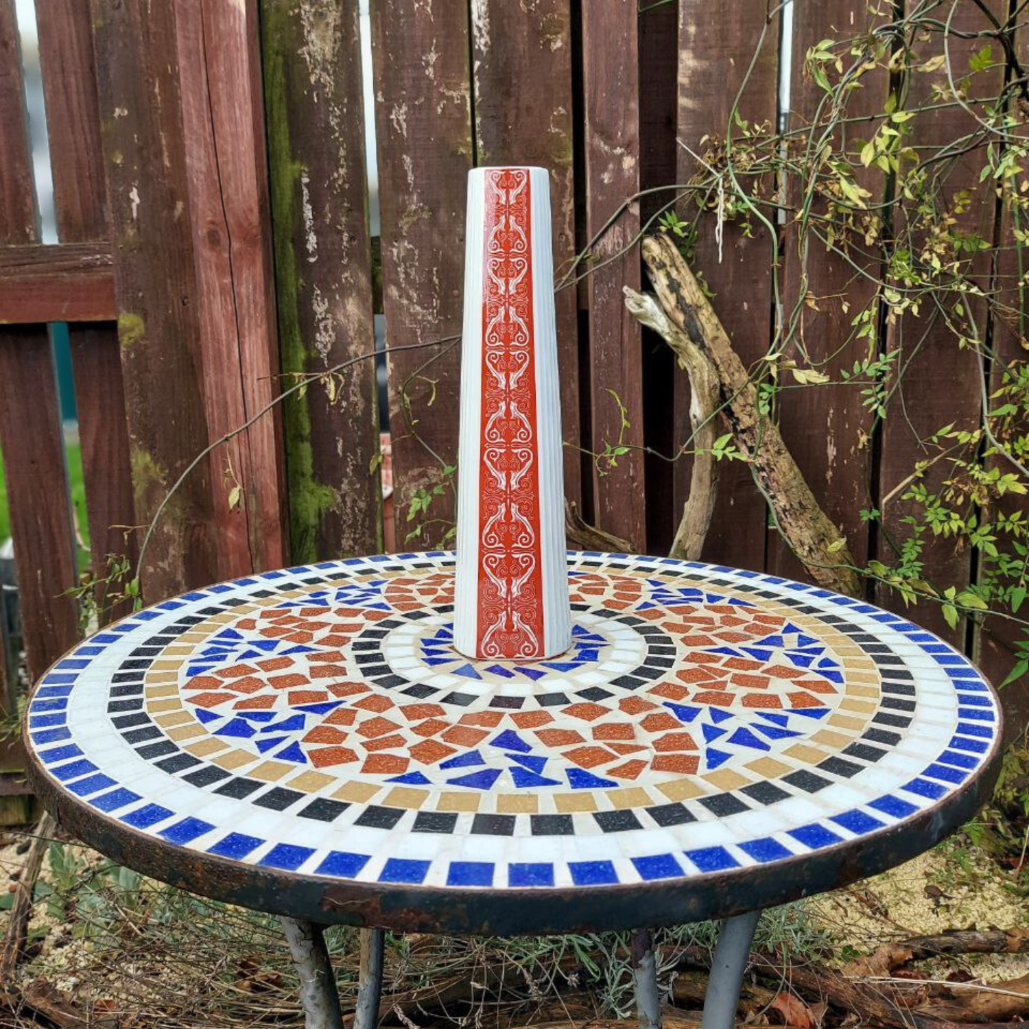 Mosaic table with a decorative vase on top, set against a wooden fence and plants.