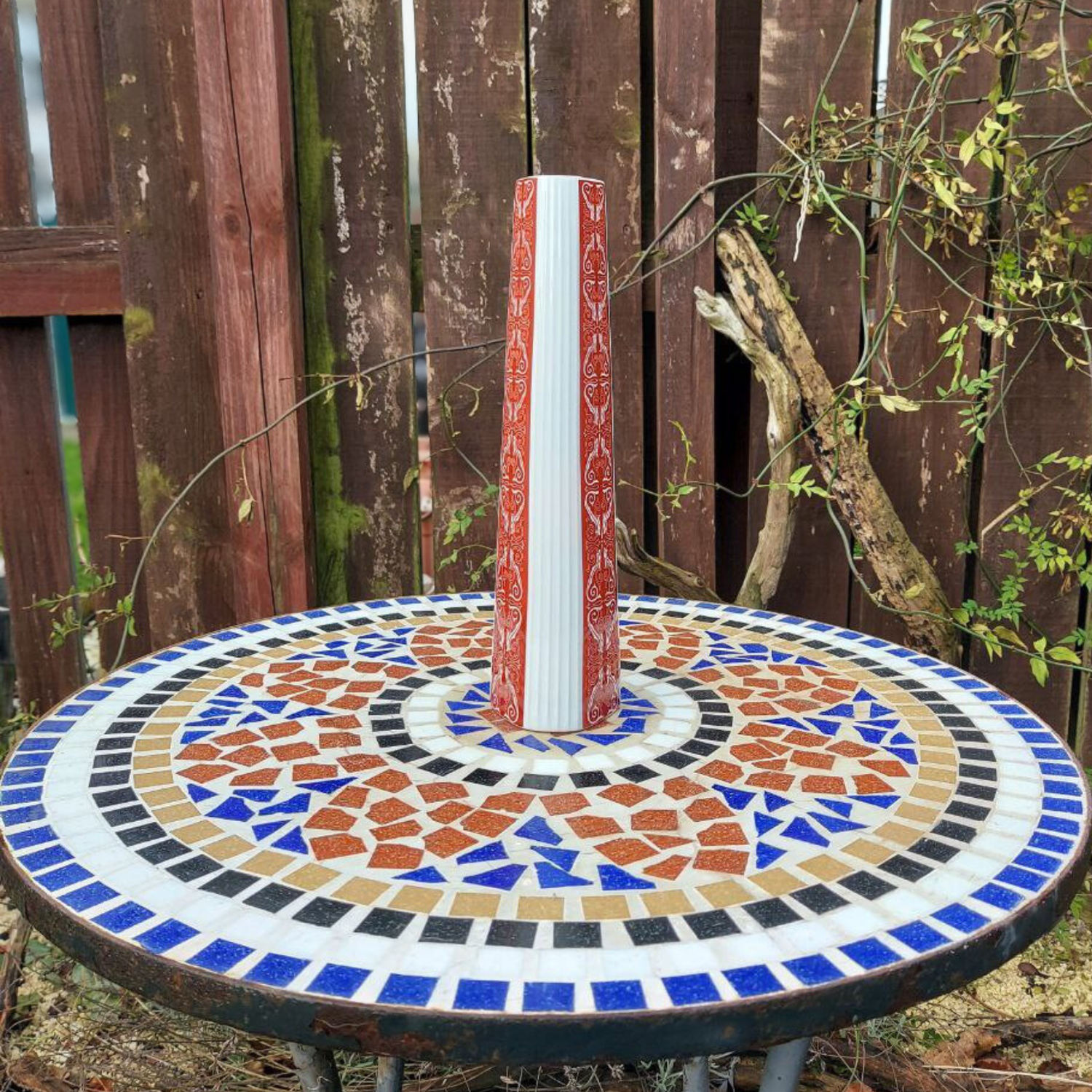 Mosaic-patterned table with a cylindrical vase on top, set against a wooden fence background.