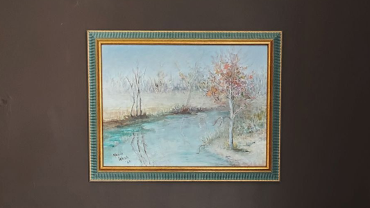 Framed painting of a landscape with a river and trees on a dark wall.