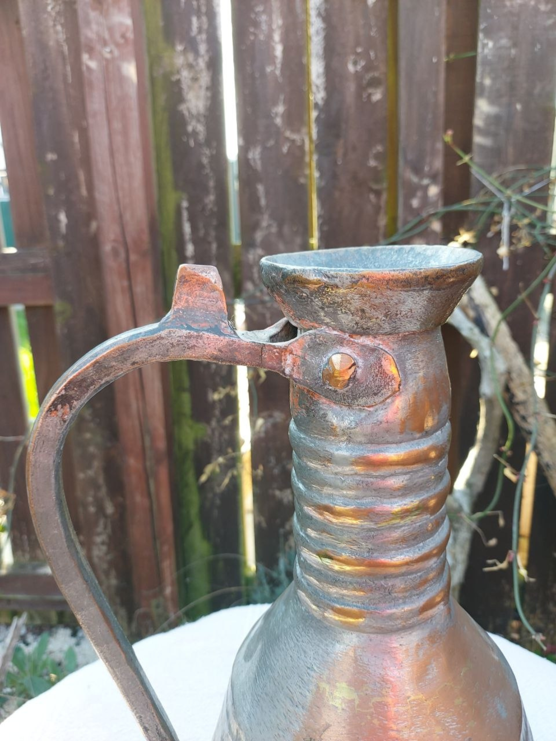 Rust-coloured copper pitcher with handle against a wooden fence background