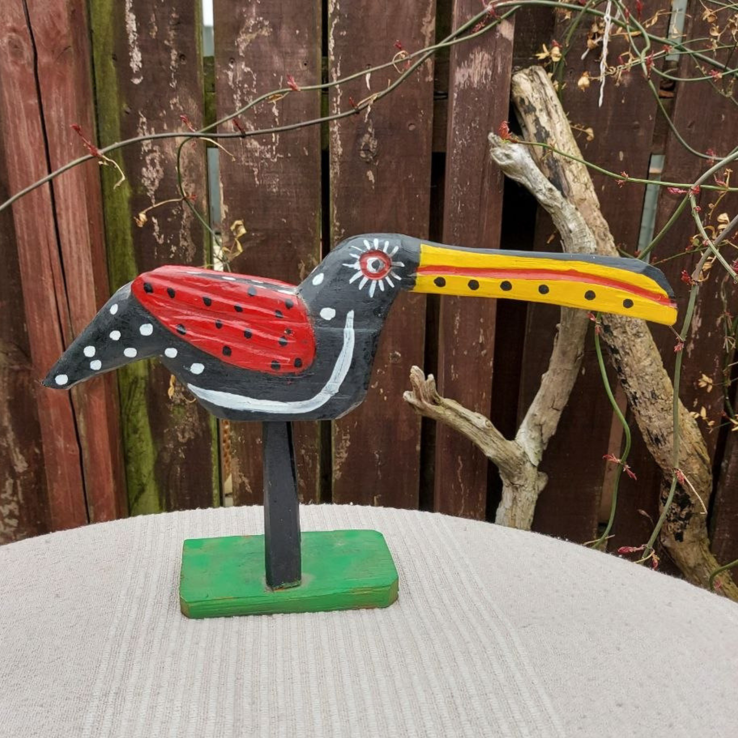 Wooden bird figurine with a red and yellow beak on a stand against a wooden fence background.