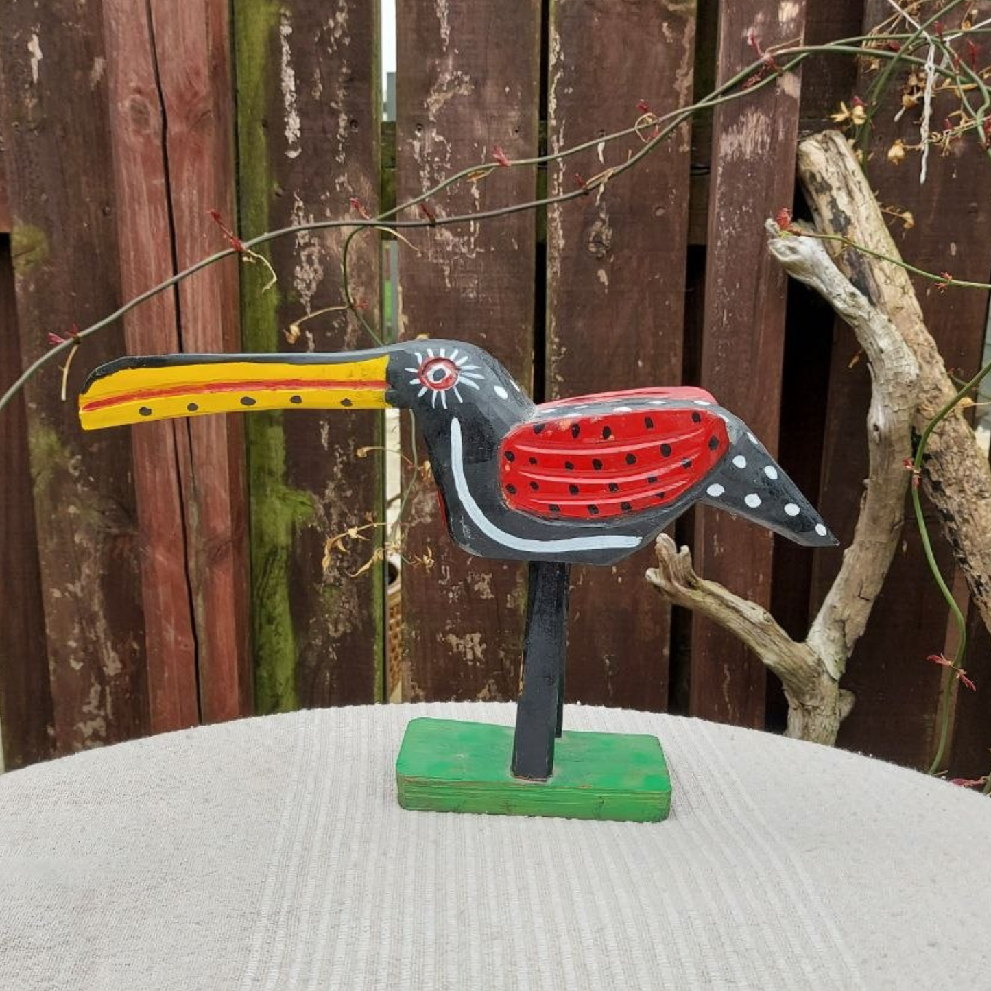 Decorative bird figurine with a long yellow beak on a stand against a wooden fence background.